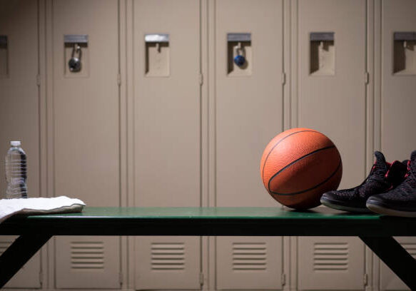 Various sports equipment on bench inside high school or college gymnasium locker room.   Items include: basketball, shoes, water, towel.